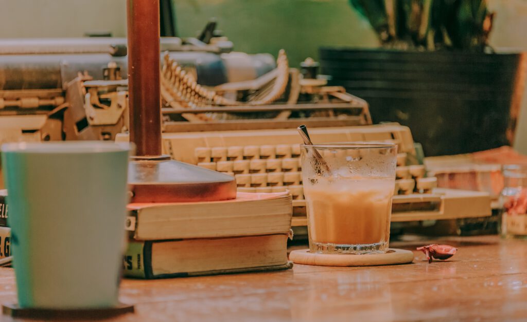 Ausschnitt eines Schreibtischs mit einer Schreibmaschine, einem Bücherstapel und einem halbvollen Getränkeglas- und Tassen