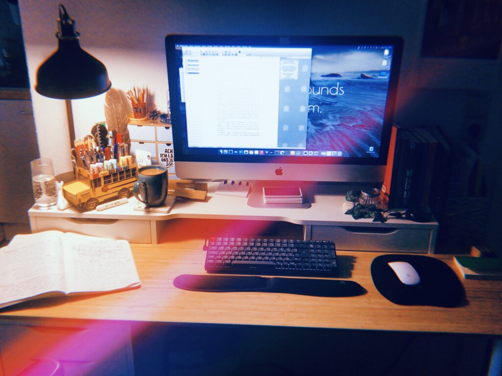 Schreibtisch mit offenem Notizheft, einer brennenden Schreibtischlampe und einem großen Computermonitor, auf dem ein offenes Textdokument zu sehen ist. Dezente Unordnung und eine Tasse Kaffee.