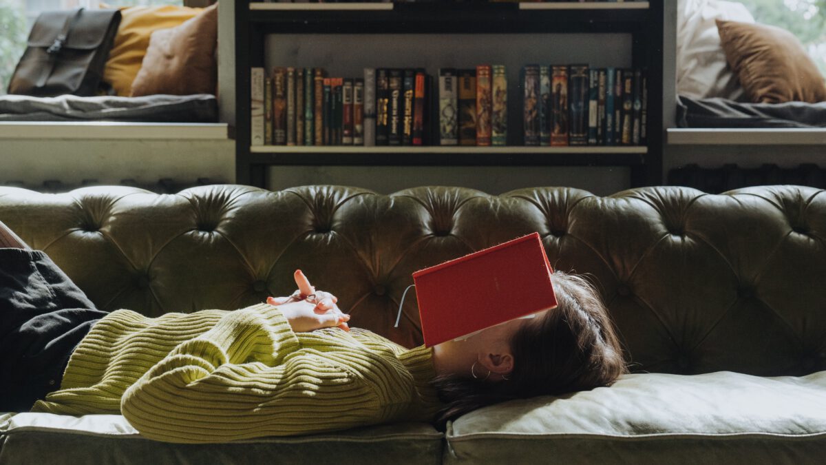 Eine Frau liegt mit vor der Brust verschränkten Händen auf einem grünen Sofa, auf ihrem Gesicht liegt ein aufgeschlagenes rotes Buch. Im Hintergrund zwei Fenster und ein Bücherregal.