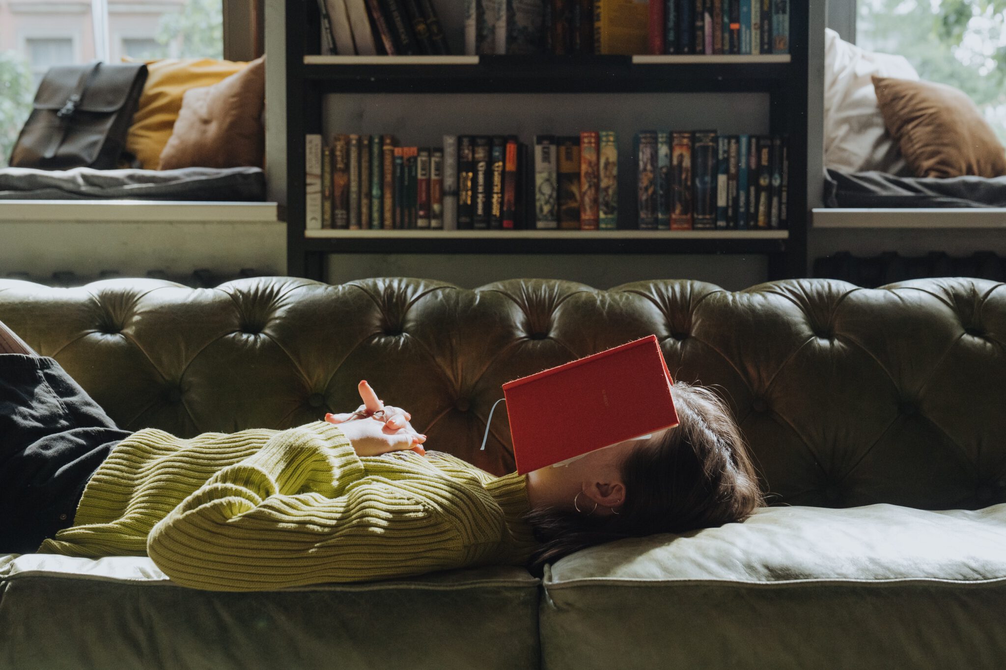 Eine Frau liegt mit vor der Brust verschränkten Händen auf einem grünen Sofa, auf ihrem Gesicht liegt ein aufgeschlagenes rotes Buch. Im Hintergrund zwei Fenster und ein Bücherregal.