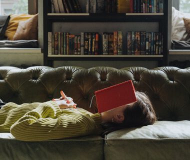 Eine Frau liegt mit vor der Brust verschränkten Händen auf einem grünen Sofa, auf ihrem Gesicht liegt ein aufgeschlagenes rotes Buch. Im Hintergrund zwei Fenster und ein Bücherregal.