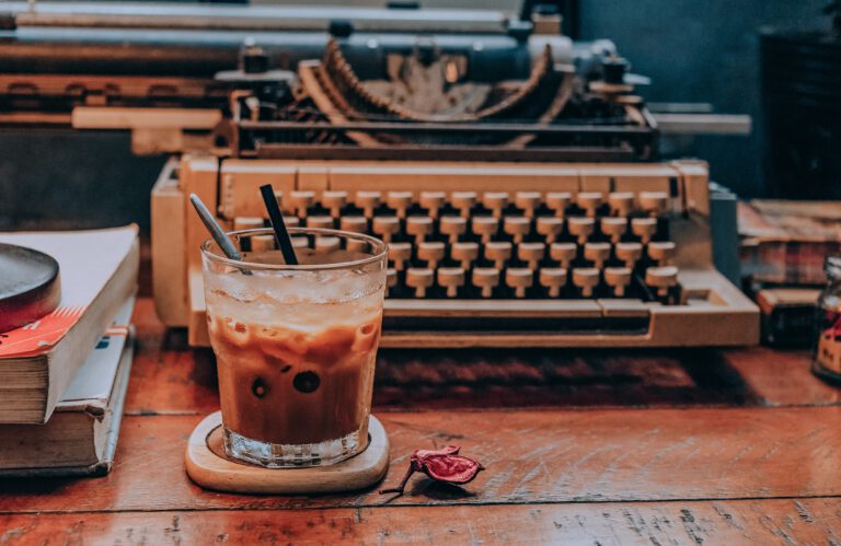 Zerkratzter unordentlicher Schreibtisch mit einer alten Schreibmaschine und einem Glas Iced Coffee auf einem hölzernen Untersetzer. Daneben liegt ein Stapel Bücher.