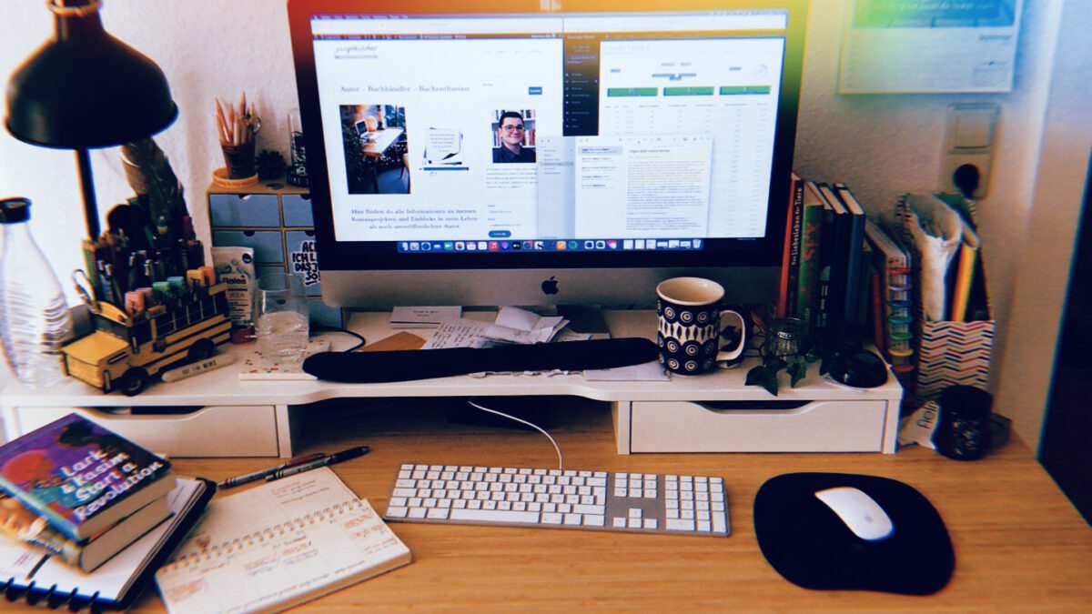 Leicht chaotischer Schreibtisch mit herumliegenden Büchern und mehreren offenen Dokumenten auf dem Computer