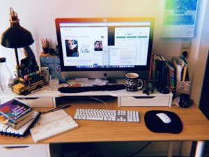 Leicht chaotischer Schreibtisch mit herumliegenden Büchern und mehreren offenen Dokumenten auf dem Computer