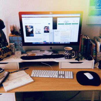 Leicht chaotischer Schreibtisch mit herumliegenden Büchern und mehreren offenen Dokumenten auf dem Computer