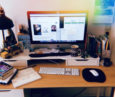 Leicht chaotischer Schreibtisch mit herumliegenden Büchern und mehreren offenen Dokumenten auf dem Computer