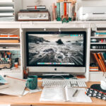 Auf dem Foto ist ein chaotischer Schreibtisch mit einem IiMac zu sehen, auf dem viele Notizzettel, Hefte und Stifte durcheinander liegen.