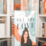 Das Taschenbuch von "All the Love – Alles anders als gedacht" steht auf einem kleinen Tisch vor einem Bücheregal