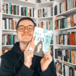 Ein Lächelnder Mann steht vor einem Bücherregal und grinst in die Kamera. Er hält das Taschenbuch von "All the Love – Alles anders als gedacht" von Ely Junge an seine Wange.